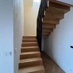 Nowoczesne drewniane schody wiszące z czarną balustradą w jasnym wnętrzu, ściany wykończone gładzią w odcieniach bieli i beżu, parkiet ułożony we wzór jodełki.