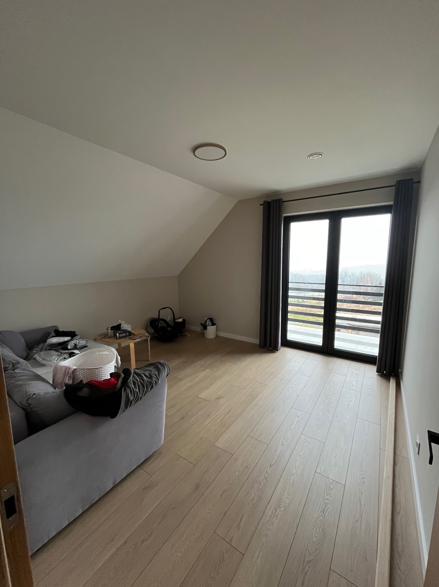 Poddasze po remoncie z jasną podłogą panelową, szarą sofą i dużymi drzwiami balkonowymi z widokiem na zewnątrz.