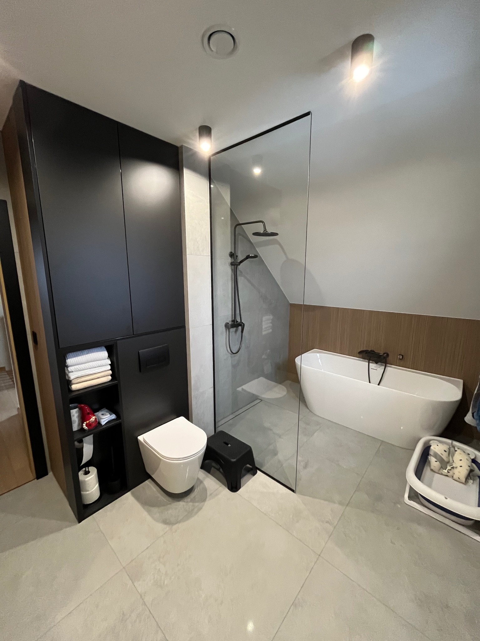 Nowoczesna łazienka na poddaszu z kabiną prysznicową typu walk-in, wolnostojącą wanną, czarnymi meblami i jasnymi płytkami podłogowymi.