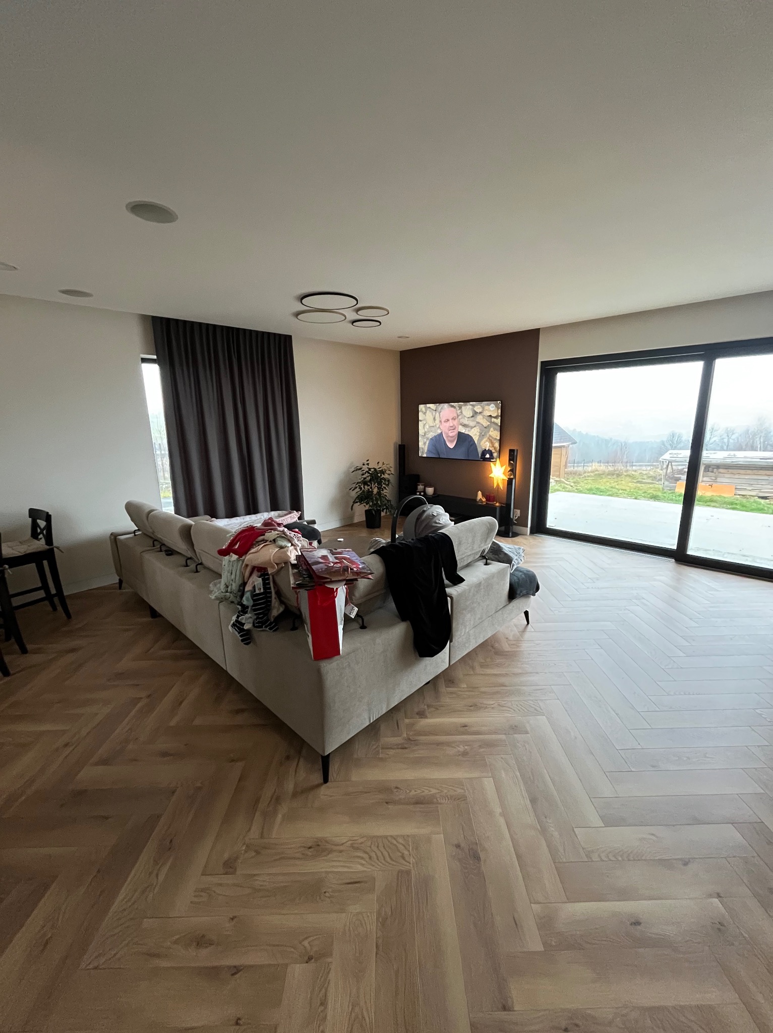 Salon z drewnianą podłogą ułożoną w jodełkę, beżowa sofa z ubraniami i prezentami, duże okno z widokiem na zewnątrz, telewizor na brązowej ścianie.