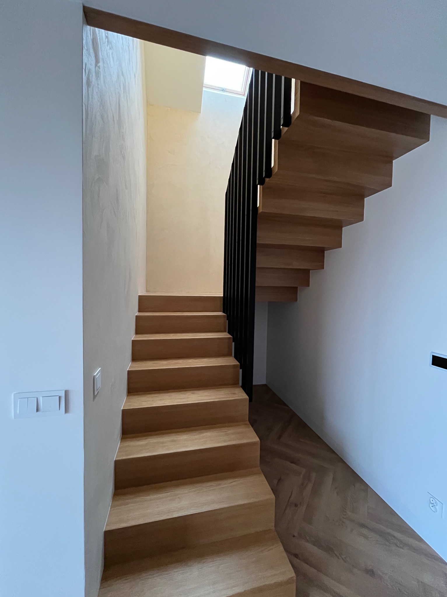 Nowoczesne drewniane schody wiszące z czarną balustradą w jasnym wnętrzu, ściany wykończone gładzią w odcieniach bieli i beżu, parkiet ułożony we wzór jodełki.