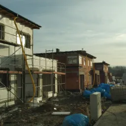 W budowie szereg domów jednorodzinnych: jeden z elewacją ocieploną styropianem i rusztowaniem, pozostałe w stanie surowym z czerwonej cegły, na pierwszym planie materiały budowlane w niebieskich...