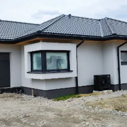 Nowoczesny dom jednorodzinny z szarą elewacją, grafitową stolarką okienną i bramą garażową, dach pokryty szarą dachówką, widoczna pompa ciepła, teren wokół domu w trakcie zagospodarowywania.