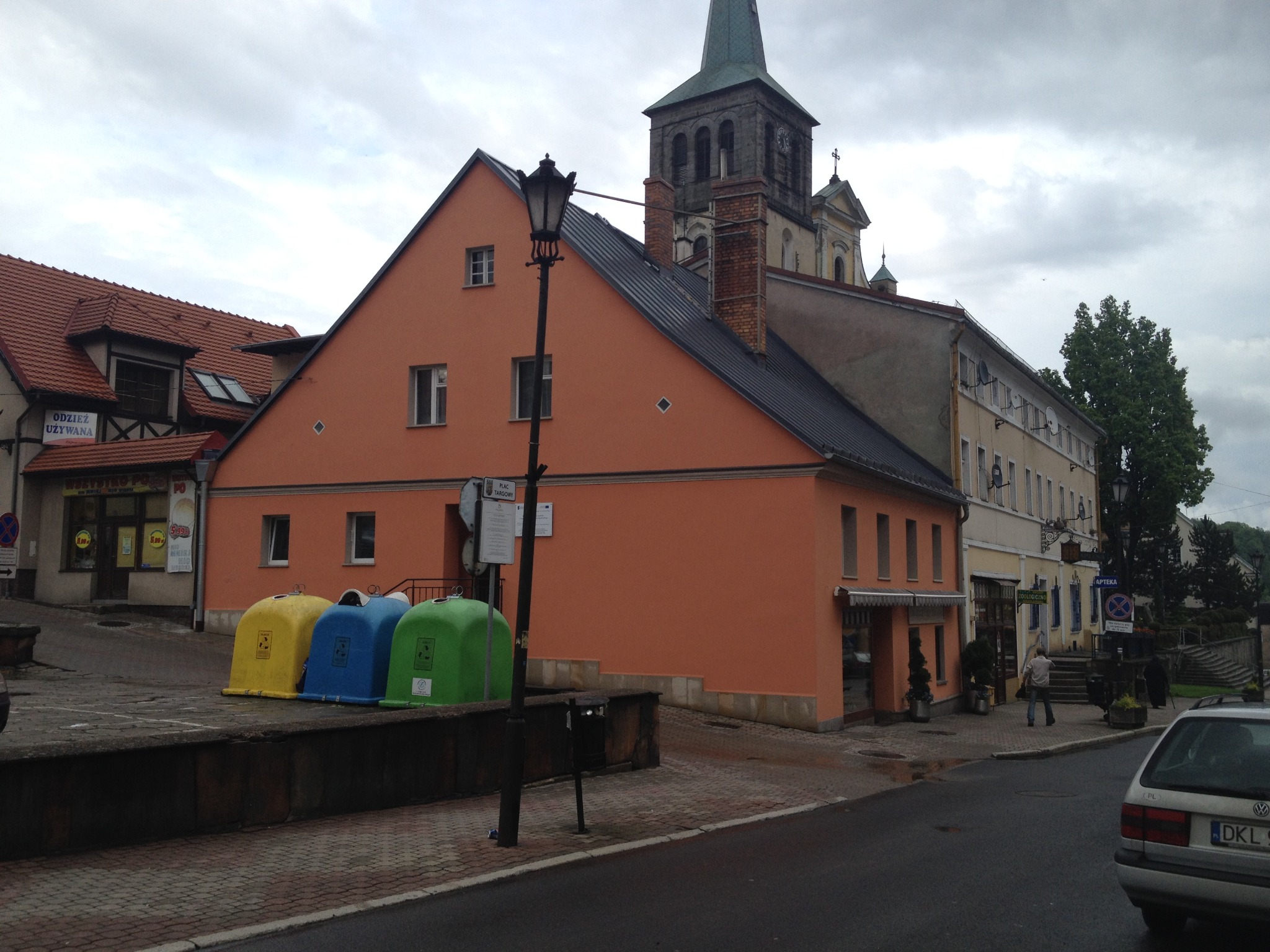 Pomarańczowy budynek z nową elewacją na tle kościoła z wieżą, widoczne kolorowe pojemniki na śmieci i fragment ulicy.