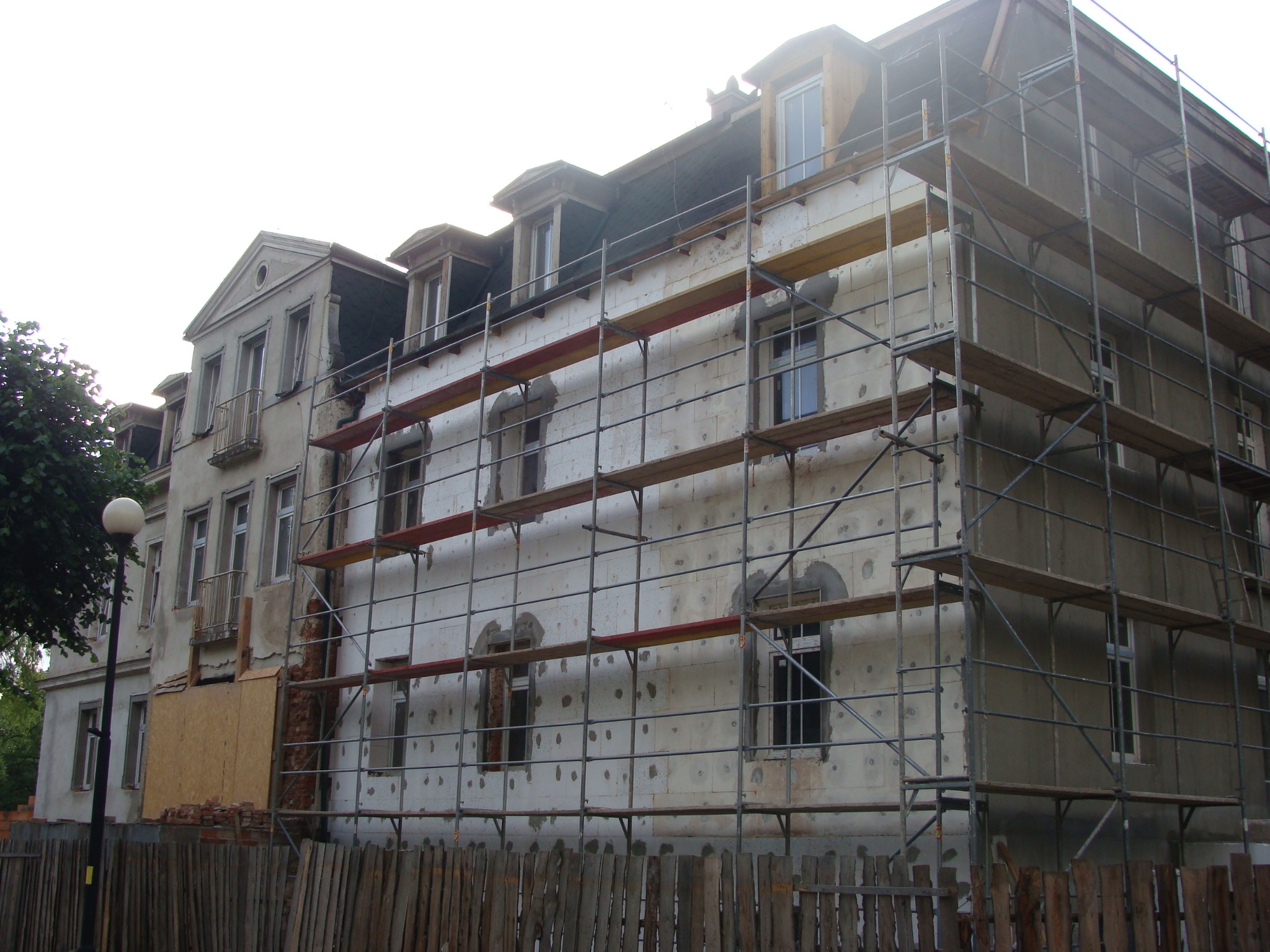 Budynek wielorodzinny w trakcie ocieplania elewacji, widoczne rusztowania, styropian i częściowo otynkowana ściana. Fragment ściany zakryty płytą OSB i sterta cegieł przy ogrodzeniu.