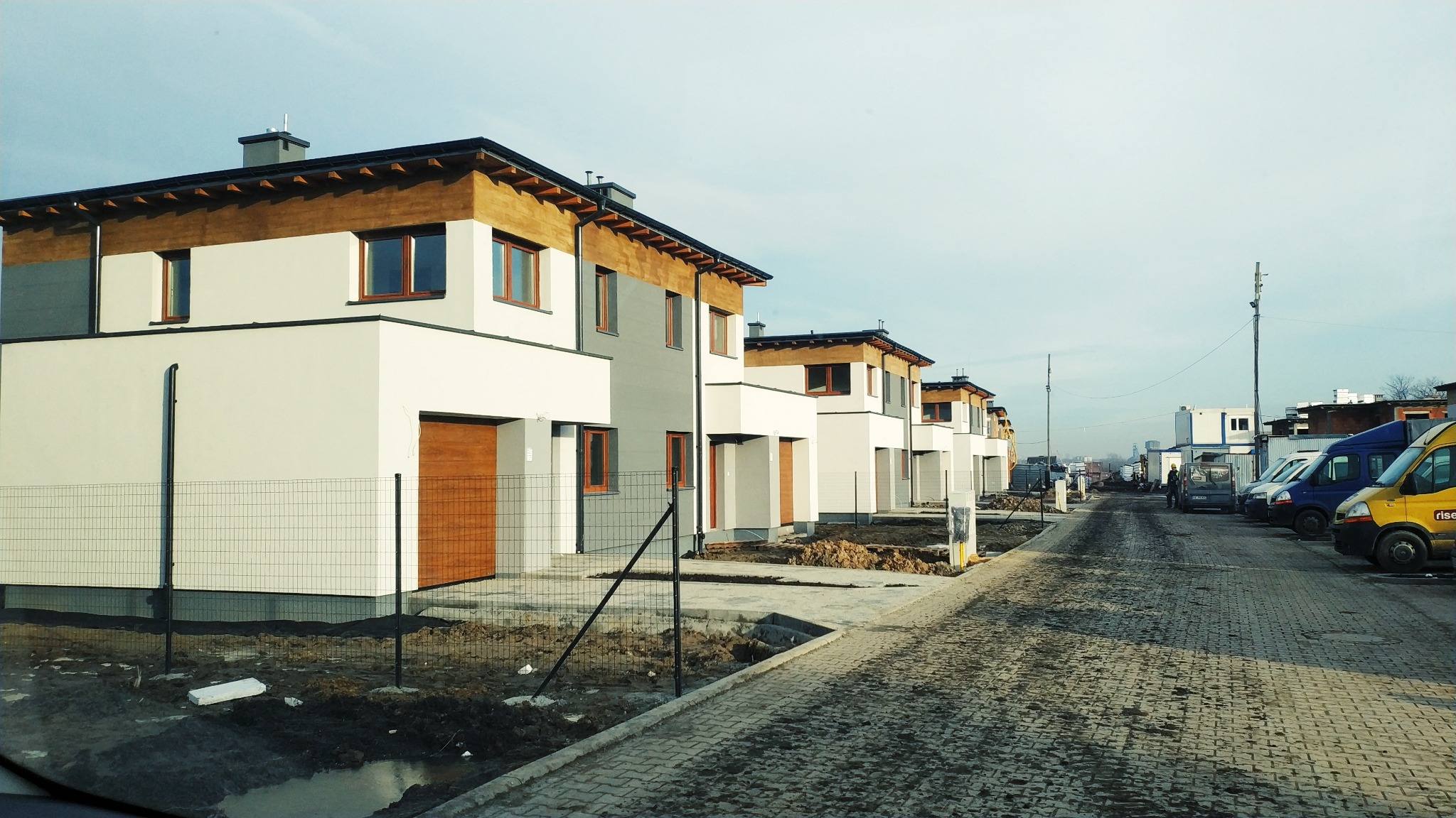 Rząd nowo wybudowanych, dwukondygnacyjnych domów szeregowych z jasną elewacją i drewnianymi akcentami, widoczny od strony ulicy z kostki brukowej, z częściowo zagospodarowanymi działkami...