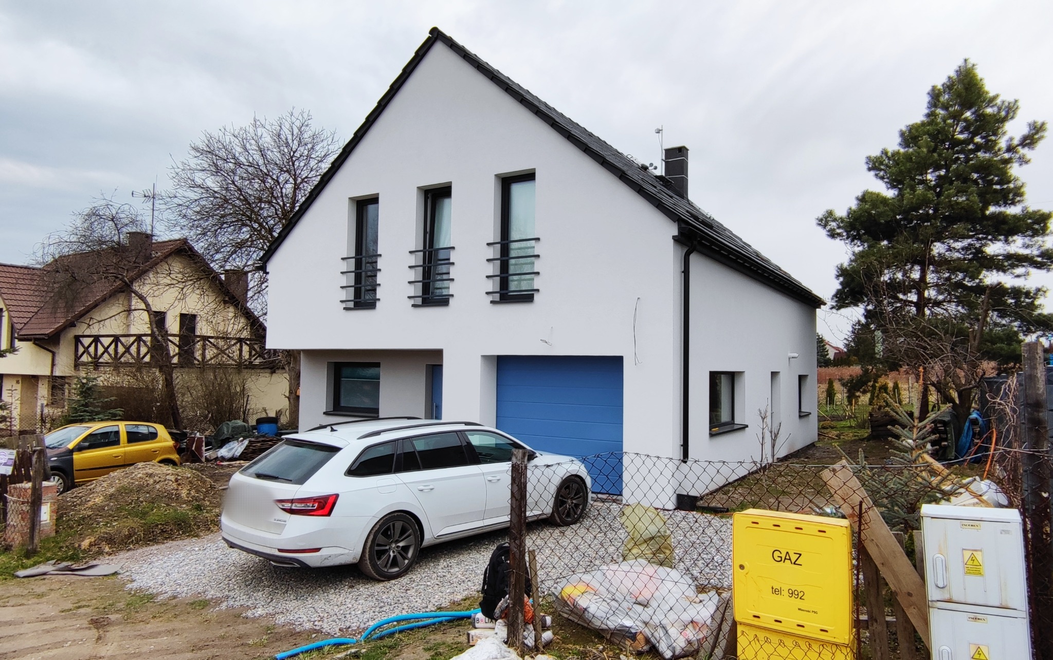 Nowoczesny dom jednorodzinny z białą elewacją, czarnym dachem i niebieską bramą garażową; widoczny samochód osobowy i skrzynka gazowa na posesji.