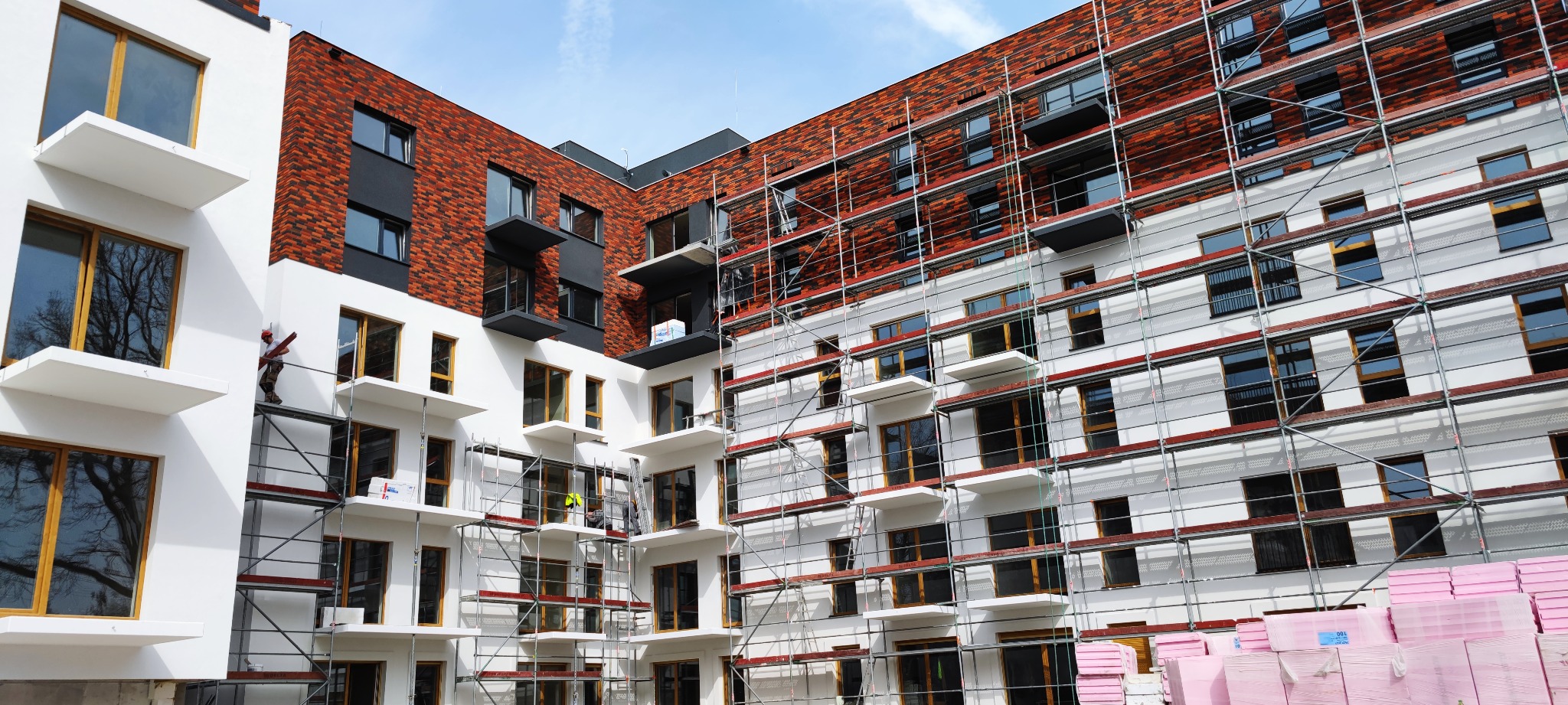 Wielopiętrowy budynek w trakcie prac elewacyjnych; widoczne rusztowania, część elewacji wykończona czerwoną cegłą, a część biała; na rusztowaniu pracownik w czerwonym kasku.