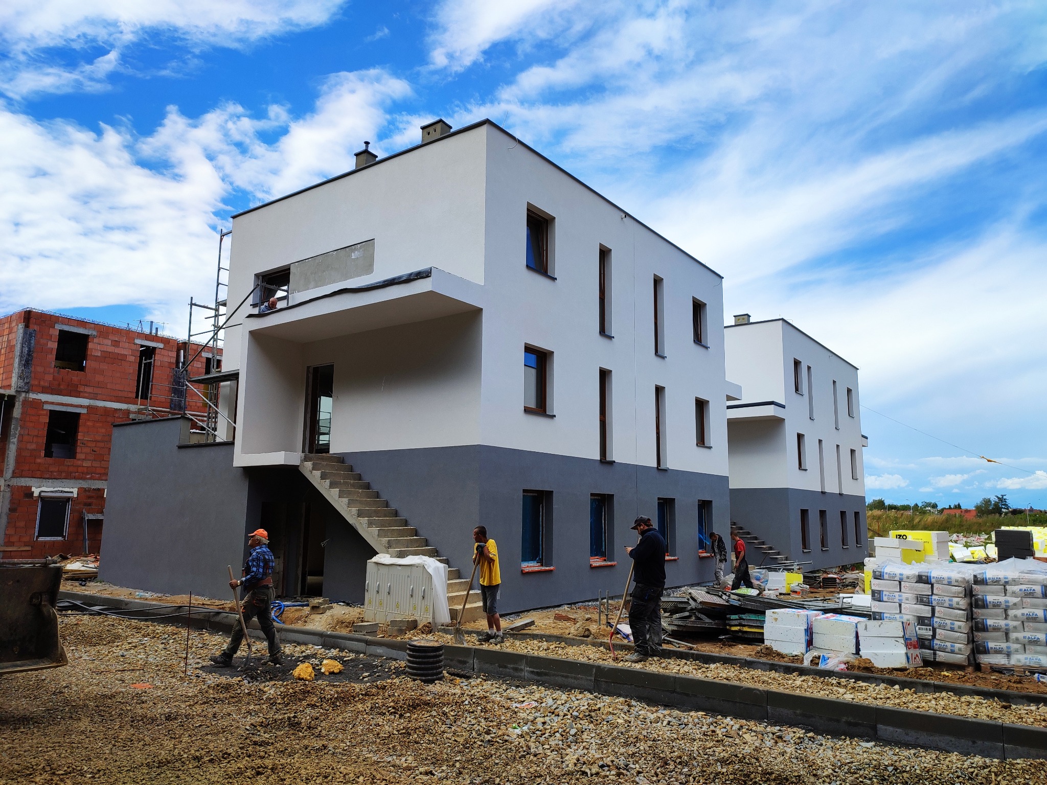 Nowoczesne domy w trakcie ocieplania elewacji, widoczni pracownicy z narzędziami, styropian i zaprawa budowlana na placu budowy pod błękitnym niebem.