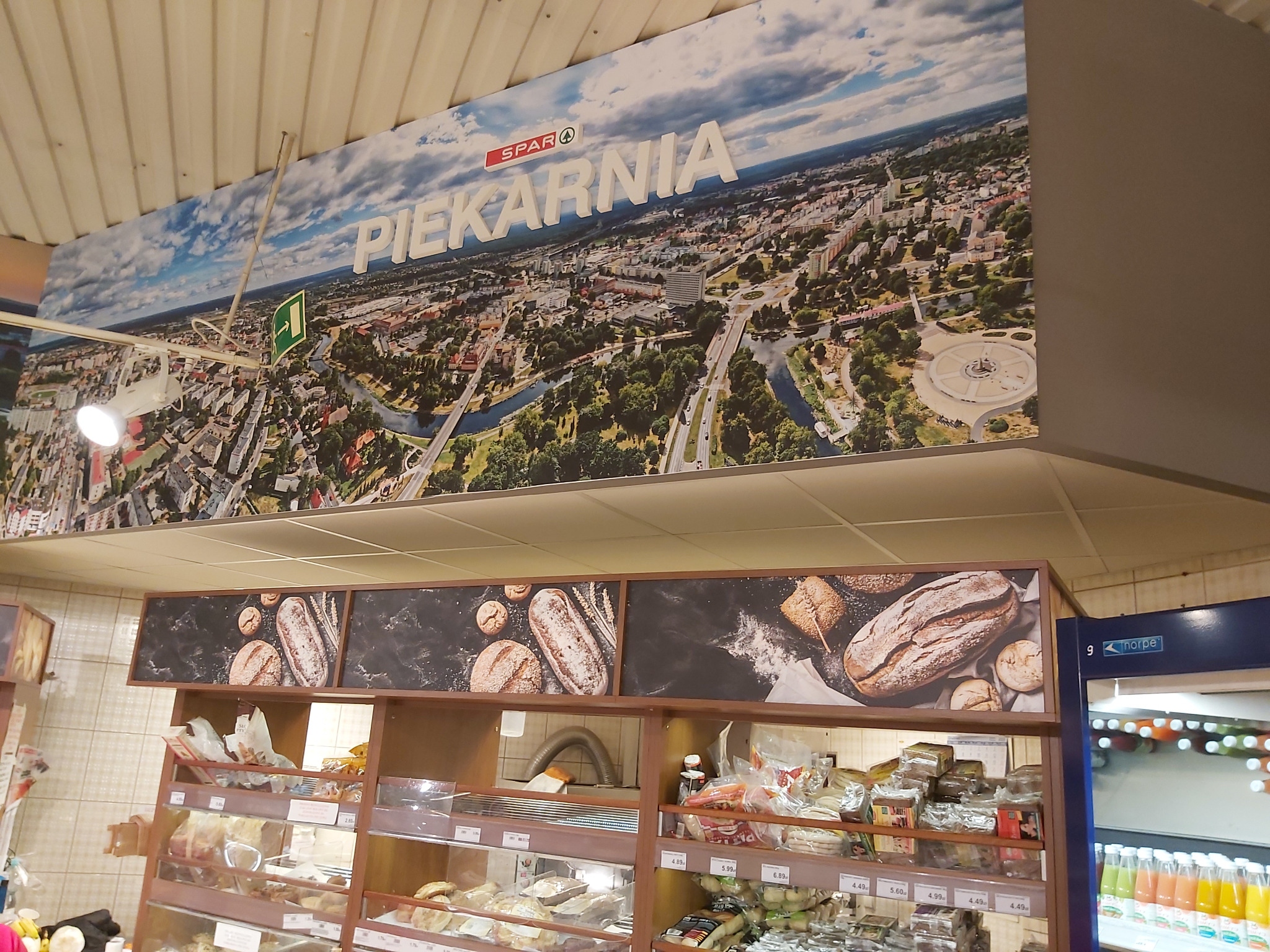 Wnętrze sklepu spożywczego z regałami pełnymi pieczywa, nad którymi wisi duży baner z napisem 'Piekarnia' i panoramą miasta widzianą z lotu ptaka.