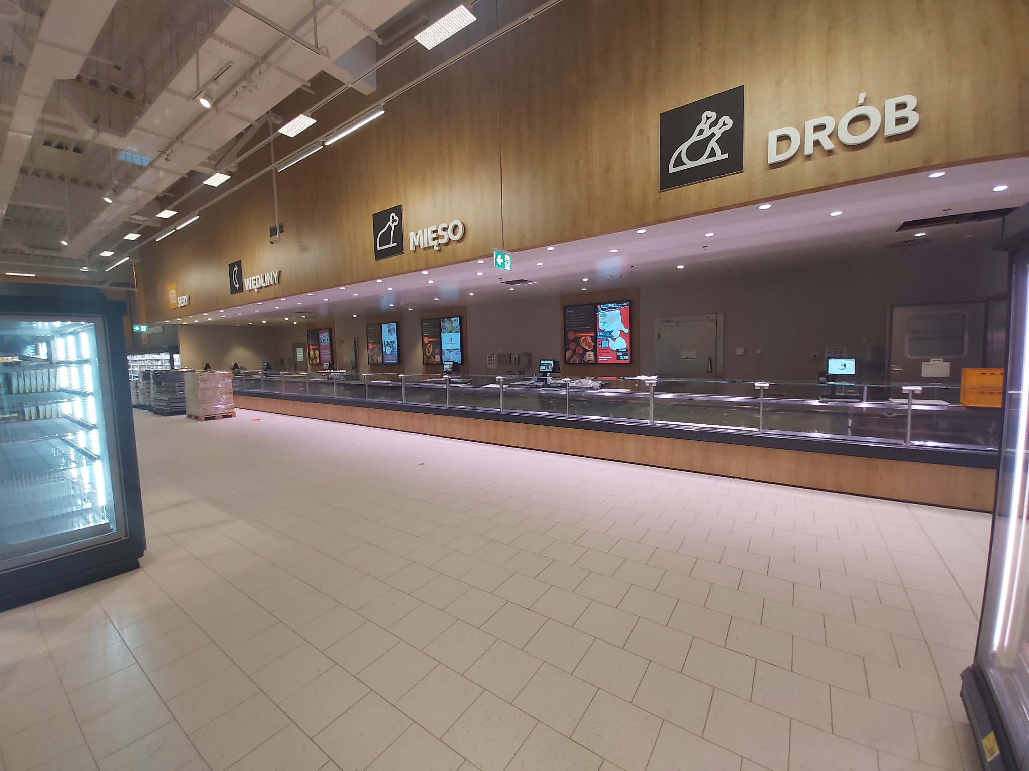 Wnętrze sklepu spożywczego z działami mięsnymi i drobiowymi, drewniane panele z logo i nazwami działów, chłodnie z produktami w tle.