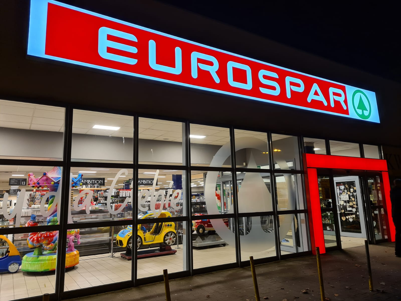 Fasada sklepu EUROSPAR z dużym czerwonym szyldem, widoczne zabawki dla dzieci za szybą i napisy na szkle.