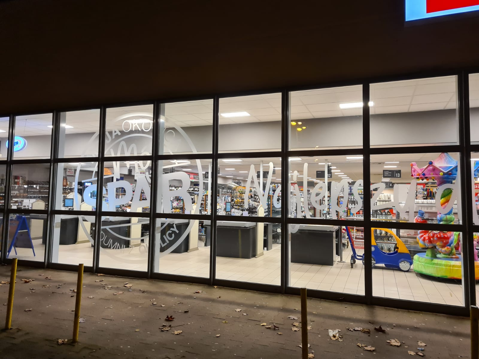 Duże okno wystawowe sklepu spożywczego z białymi napisami promocyjnymi i logo sieci. Widoczne wnętrze sklepu z kasami i towarami.