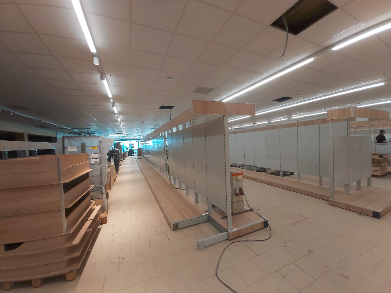 Wnętrze sklepu w trakcie montażu regałów, widoczne puste półki i materiały budowlane, oświetlenie sufitowe