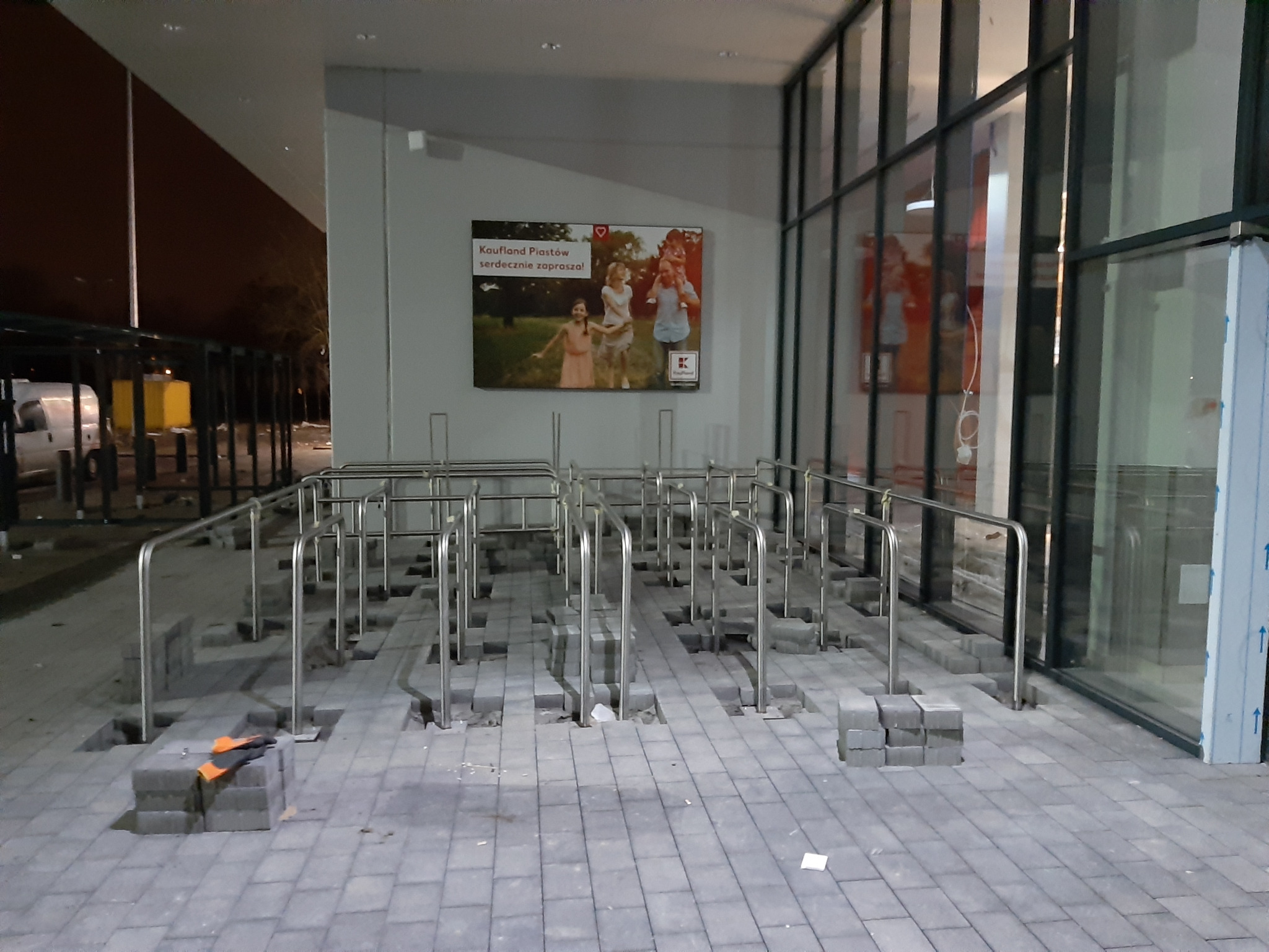 Montaż metalowych barierek i układanie kostki brukowej na zewnątrz budynku z reklamą sklepu na ścianie.