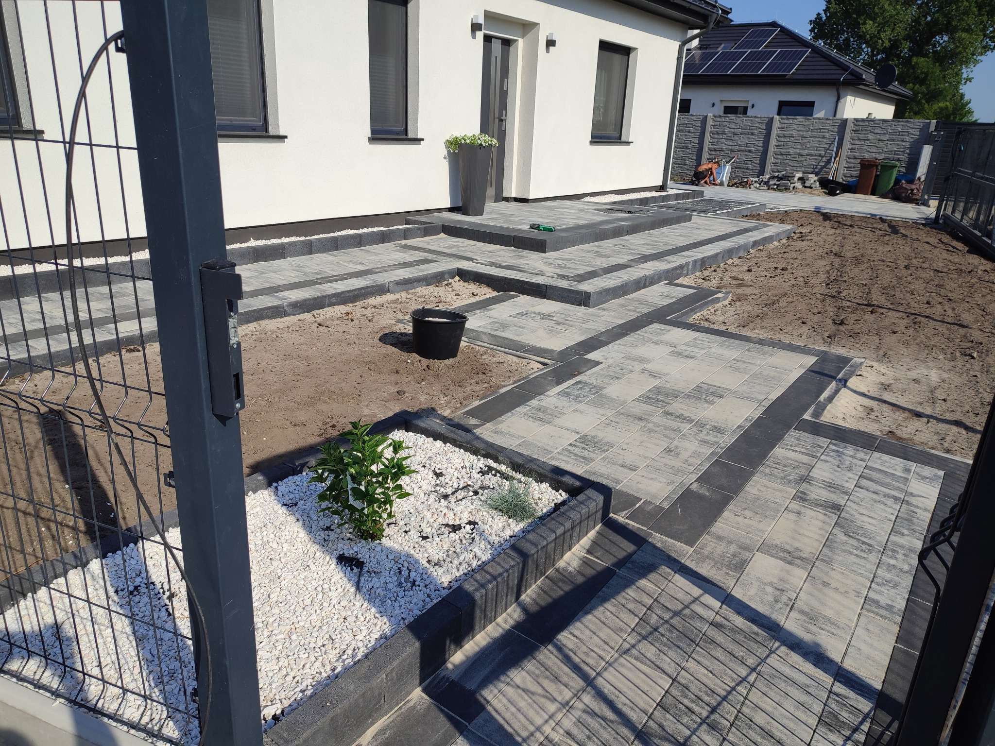 Ułożona kostka brukowa w odcieniach szarości prowadząca do domu z donicą kwiatów, z widocznym fragmentem rabaty z białym żwirem i roślinami, oraz elementem ogrodzenia w tle.