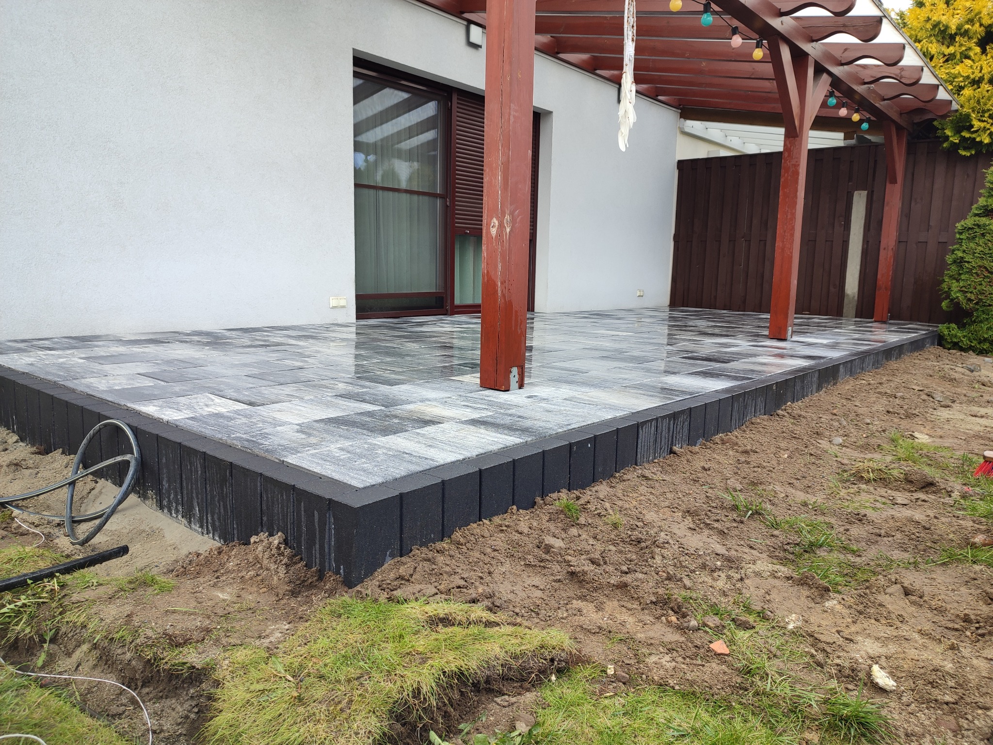 Nowo ułożony taras z kostki w szaro-czarną szachownicę, z ciemnymi obrzeżami, częściowo widoczna drewniana pergola z kolorowymi lampkami, teren wokół w trakcie prac ziemnych.