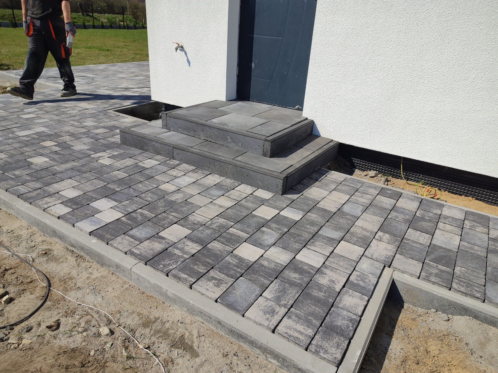 Ułożona kostka brukowa w odcieniach szarości prowadząca do schodów wejściowych domu, widoczny fragment pracownika w tle.