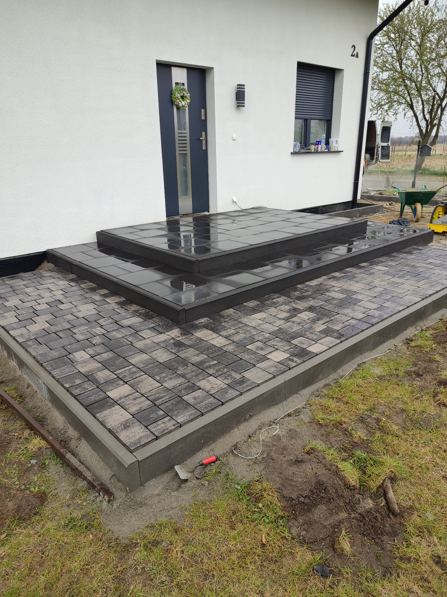 Nowo ułożony, dwustopniowy taras z kostki brukowej w odcieniach szarości, prowadzący do drzwi wejściowych domu z wieńcem. Widoczne narzędzia brukarskie i świeże obrzeże betonowe.