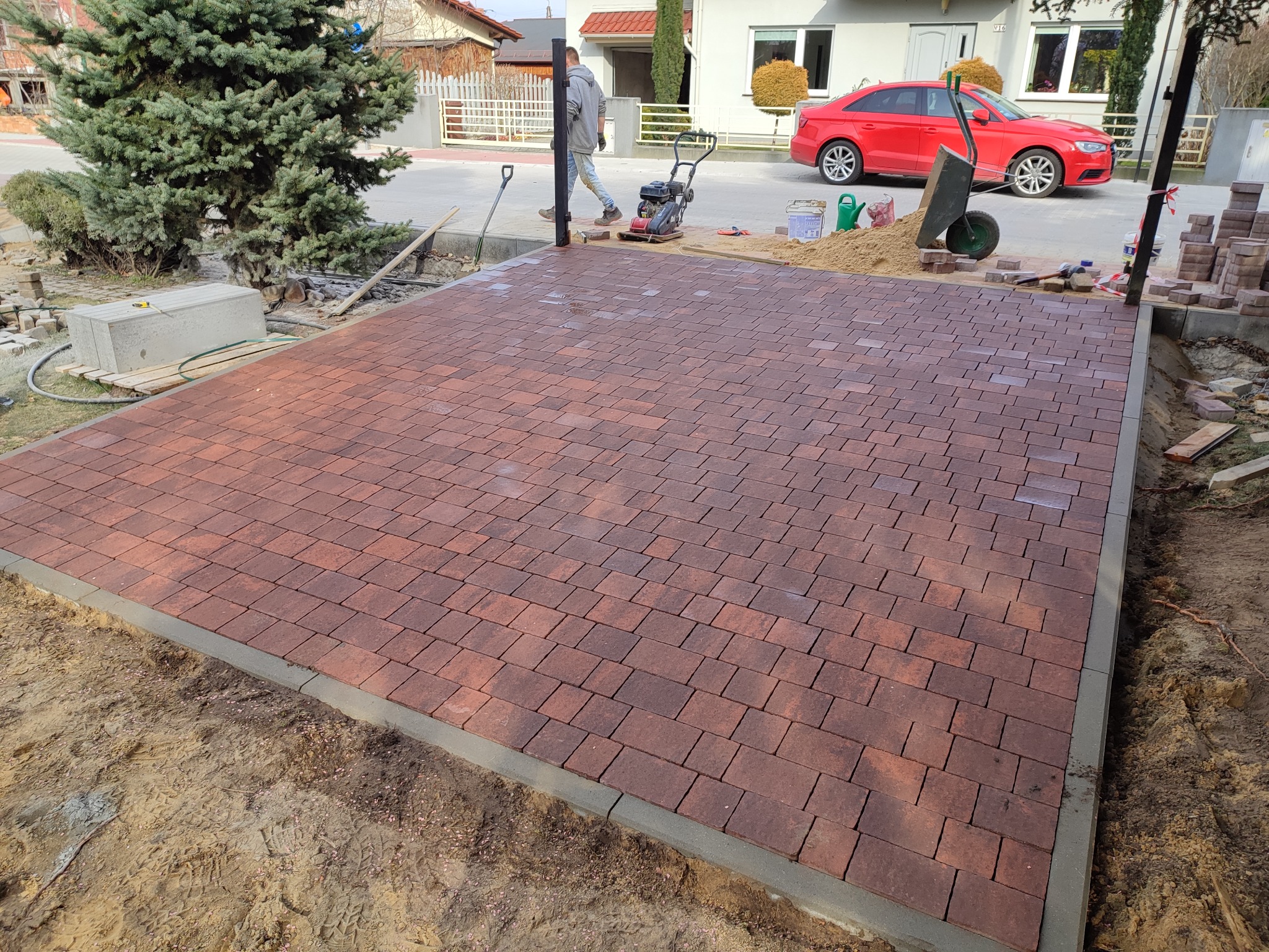 Częściowo ułożona nawierzchnia z czerwonej kostki brukowej, widoczny zagęszczarka, taczka z piaskiem, oraz przygotowane obrzeża w otoczeniu domów jednorodzinnych.