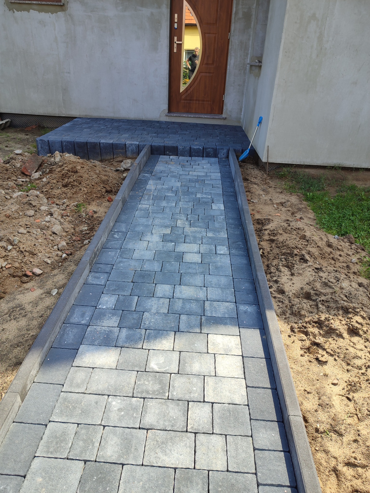 Ścieżka z szarej kostki brukowej prowadząca do drzwi wejściowych domu w trakcie układania, widoczna niebieska łopata oparta o ścianę budynku.