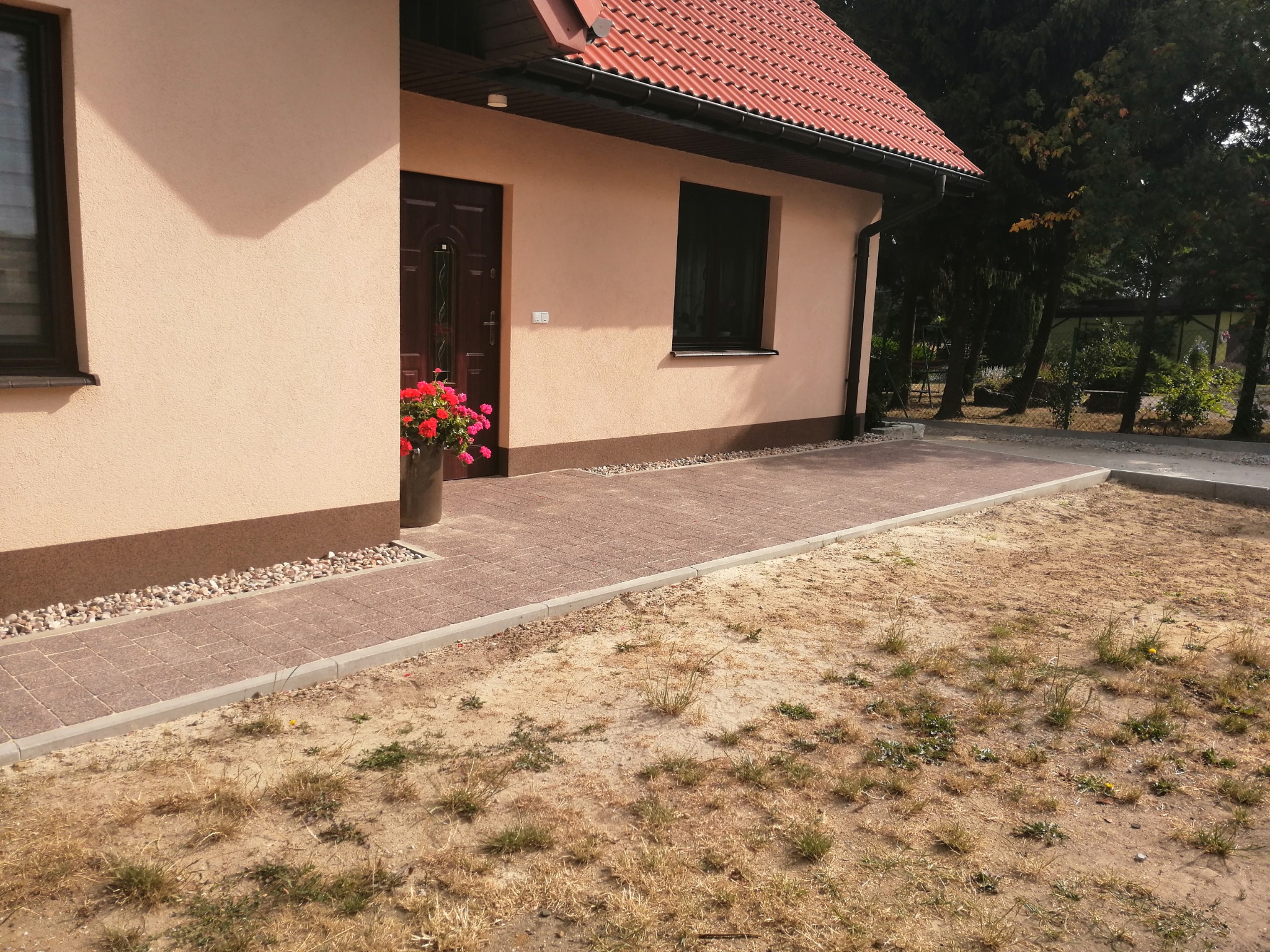 Ułożona ścieżka z kostki brukowej prowadząca do drzwi wejściowych domu, obok rabata z drobnymi kamieniami i donica z czerwonymi kwiatami.