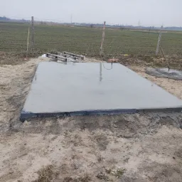 Świeżo wylana, mokra płyta betonowa na gruncie, z widocznymi śladami po narzędziach i szalunku, otoczona ziemią i trawą, w tle pole i słupy wysokiego napięcia.