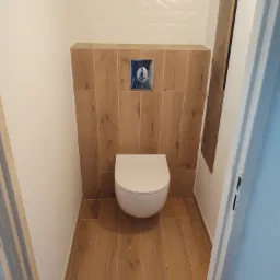 Zabudowana toaleta podtynkowa w małej łazience, wykończona płytkami imitującymi drewno, z ozdobną białą glazurą u góry i chromowanym przyciskiem spłukującym.