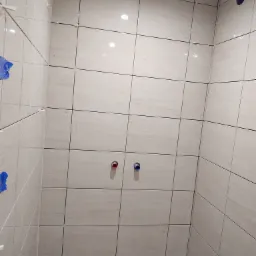 Świeżo ułożone, beżowe płytki w łazience, z krzyżykami dystansowymi w fugach i wystającymi przyłączami wodnymi z czerwonym i niebieskim oznaczeniem.