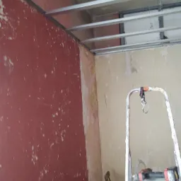 Metalowa konstrukcja sufitu podwieszanego z widocznymi profilami, ściana z resztkami farby, drabina z wiertarką na tle remontowanego pomieszczenia.