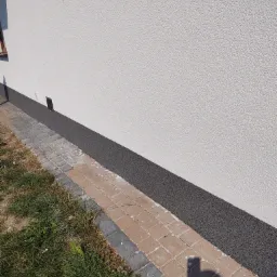 Wykończona elewacja budynku z szarą, żywiczną listwą przypodłogową i kostką brukową na obrzeżu, widok z poziomu gruntu.