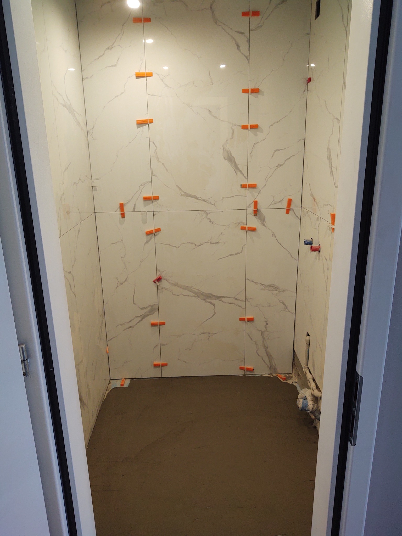 Łazienka w trakcie remontu: białe płytki imitujące marmur na ścianach, pomarańczowe krzyżyki dystansowe, wylewka na podłodze, widoczne rury instalacji wodnej.