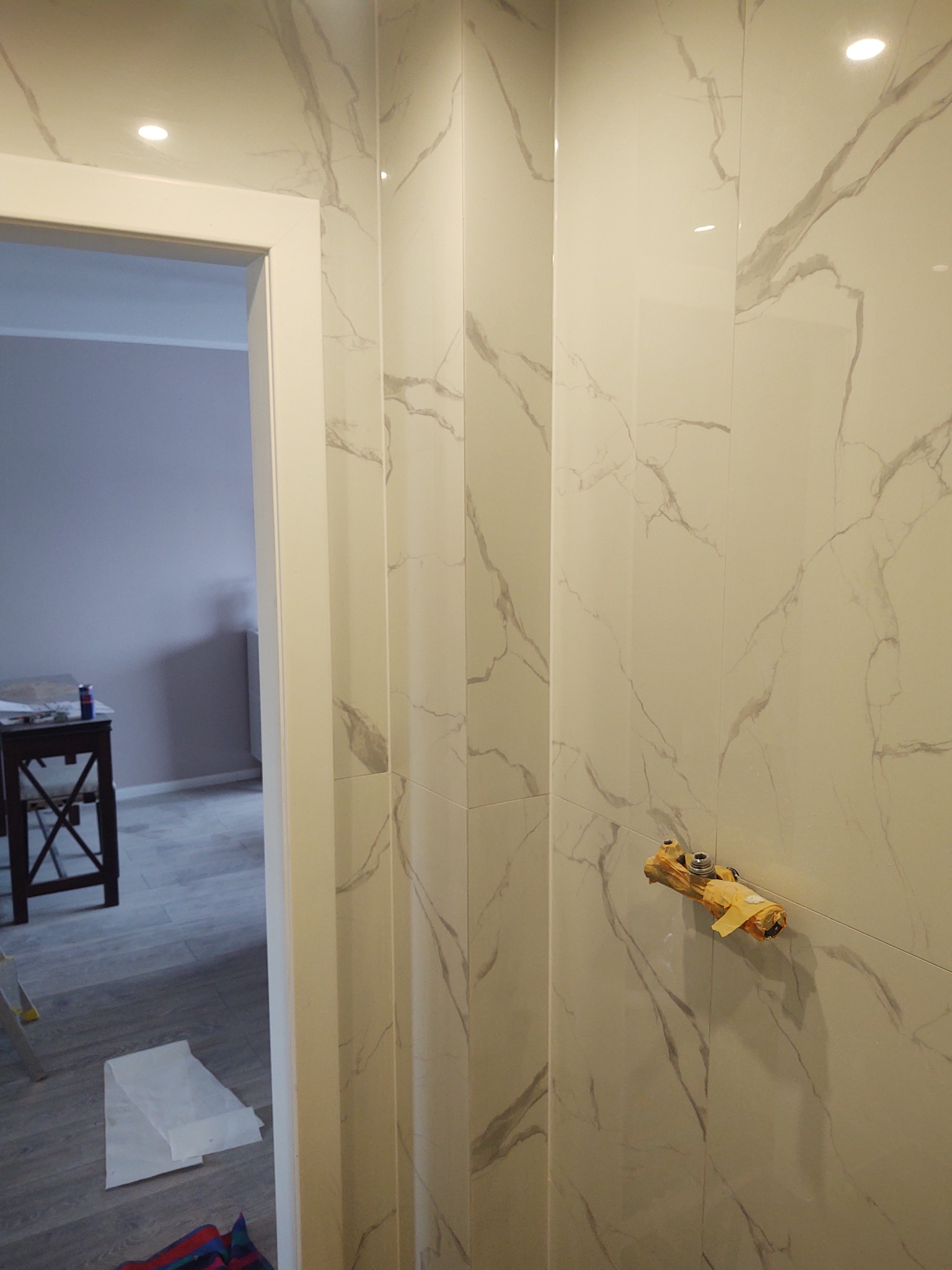 Świeżo wykończona łazienka z białymi płytkami imitującymi marmur, widoczne zabezpieczone zawory wodne, fragment pokoju z ciemnym stolikiem w tle.