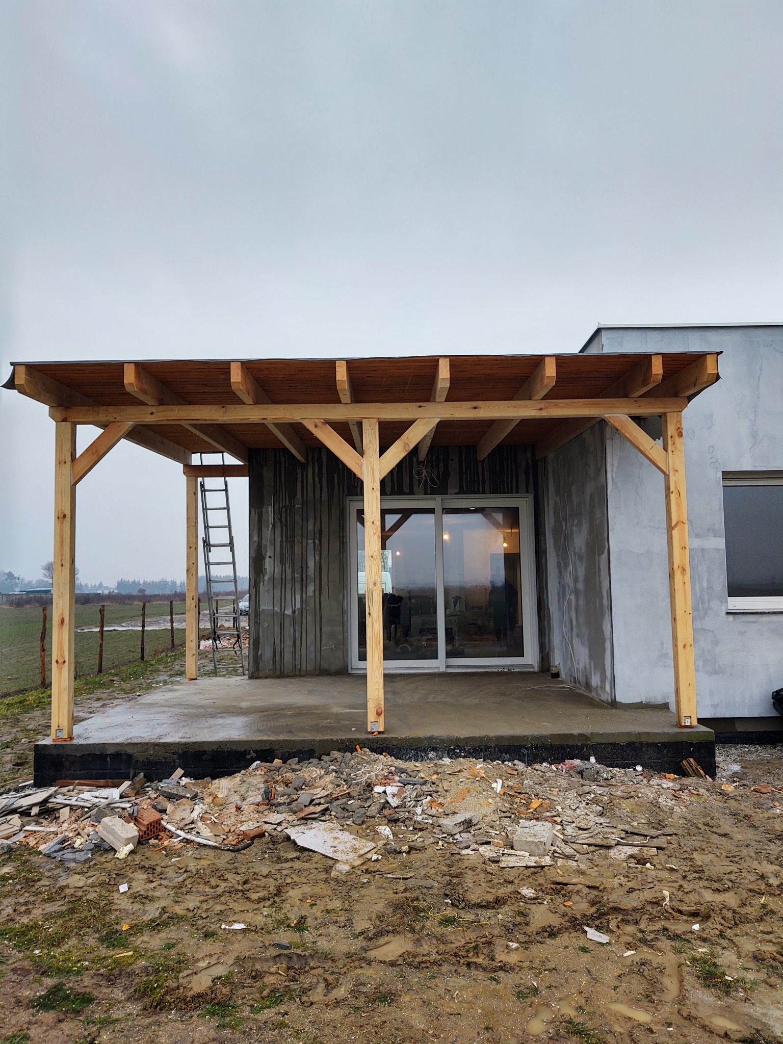 Drewniana konstrukcja zadaszenia tarasu w trakcie budowy, z widoczną drabiną opartą o ścianę budynku i gruzem budowlanym na błotnistym podłożu.