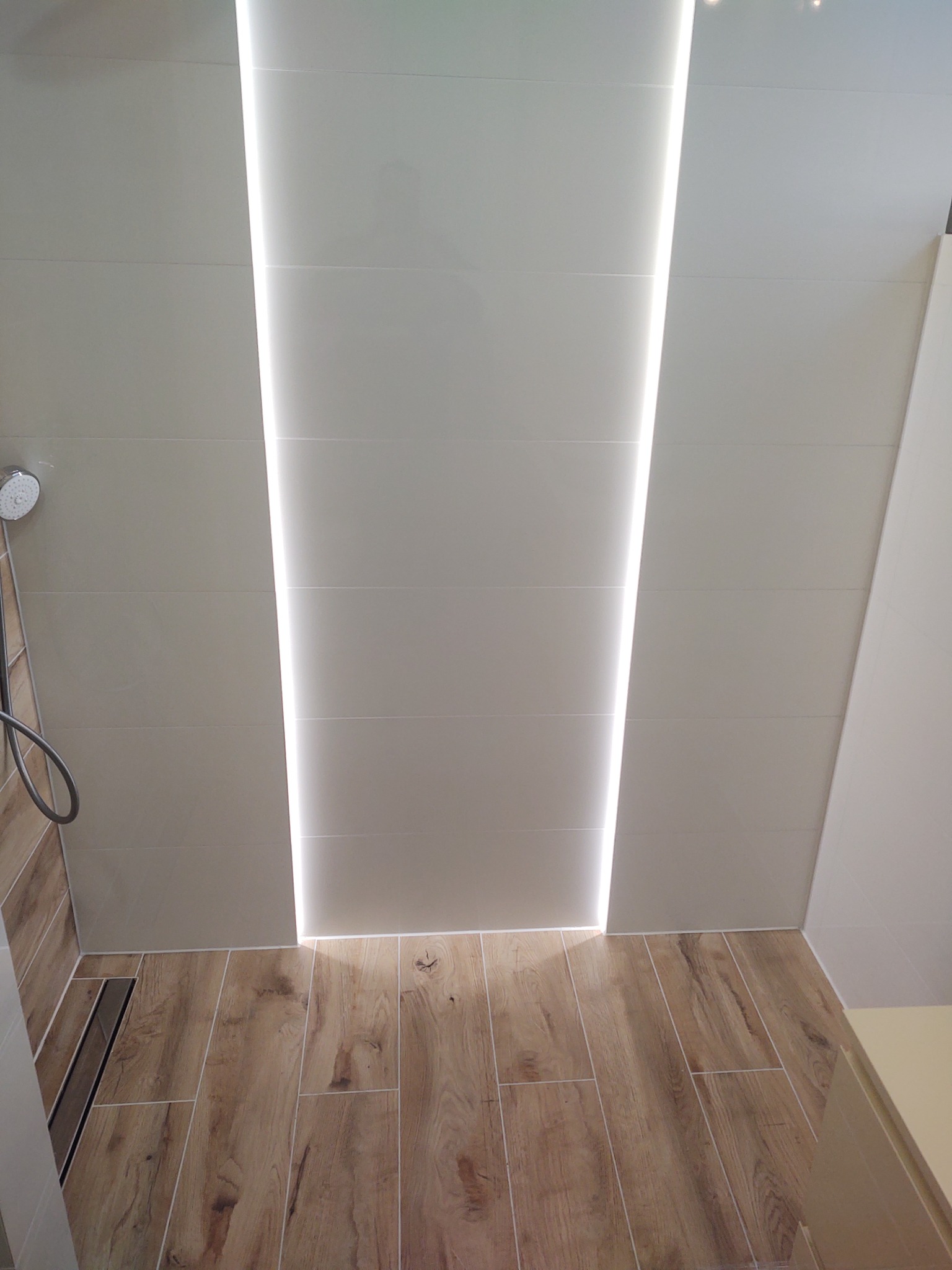 Wykończona łazienka z drewnopodobną podłogą, beżowymi płytkami na ścianach i wbudowanym oświetleniem LED w suficie, widoczny odpływ liniowy.