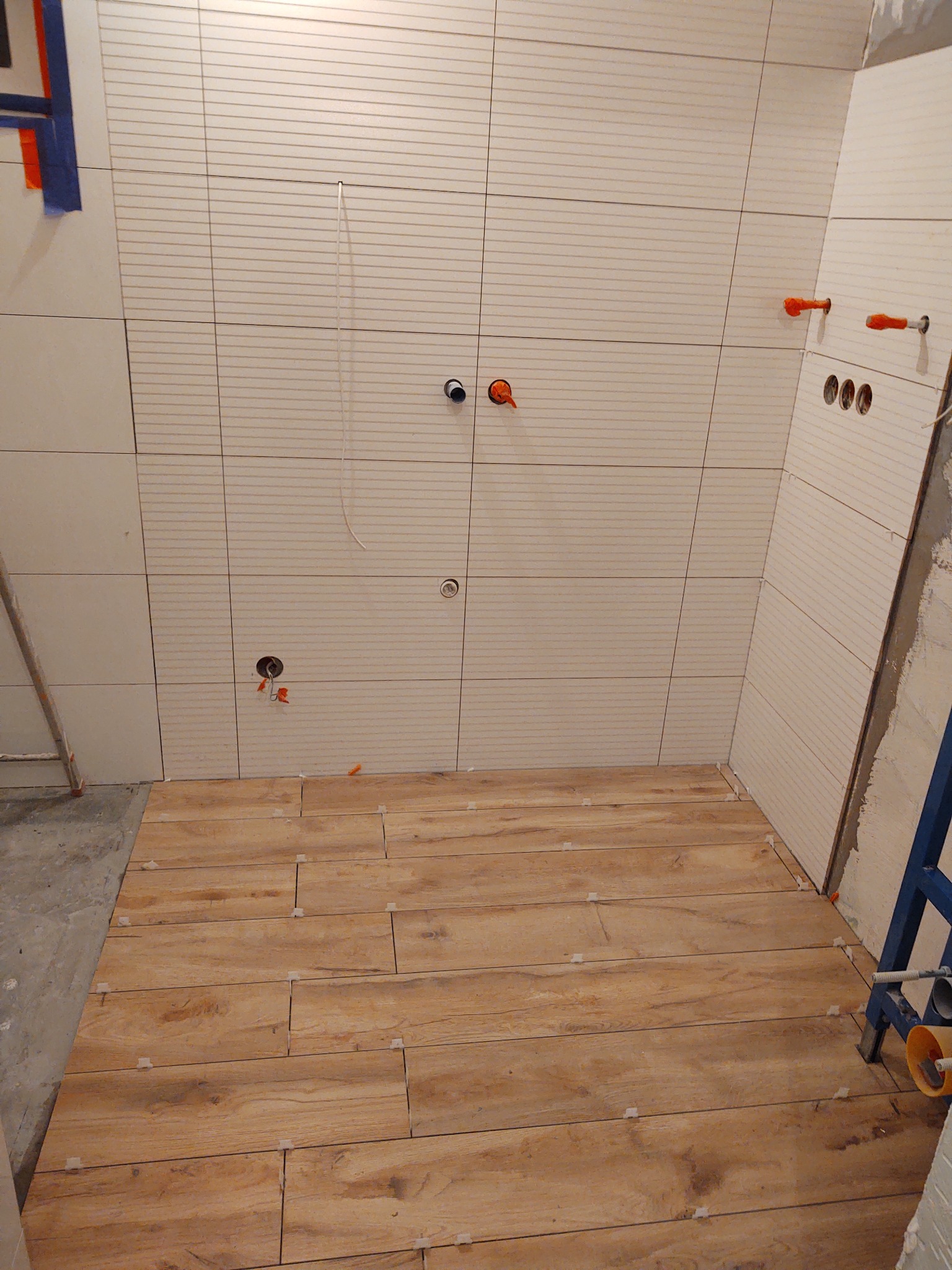 Wnętrze łazienki w trakcie prac wykończeniowych: ułożone płytki imitujące drewno na podłodze, jasne kafelki z poziomymi frezami na ścianach, widoczne otwory na armaturę i rurki, oraz plastikowe...