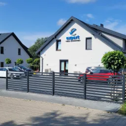Dwukondygnacyjny budynek biurowy z szarą dachówką i białymi ścianami, z niebieskim logo 'expert' nad wejściem, ogrodzony szarą, poziomą metalową barierką, z zaparkowanymi samochodami i drzewami w tle.