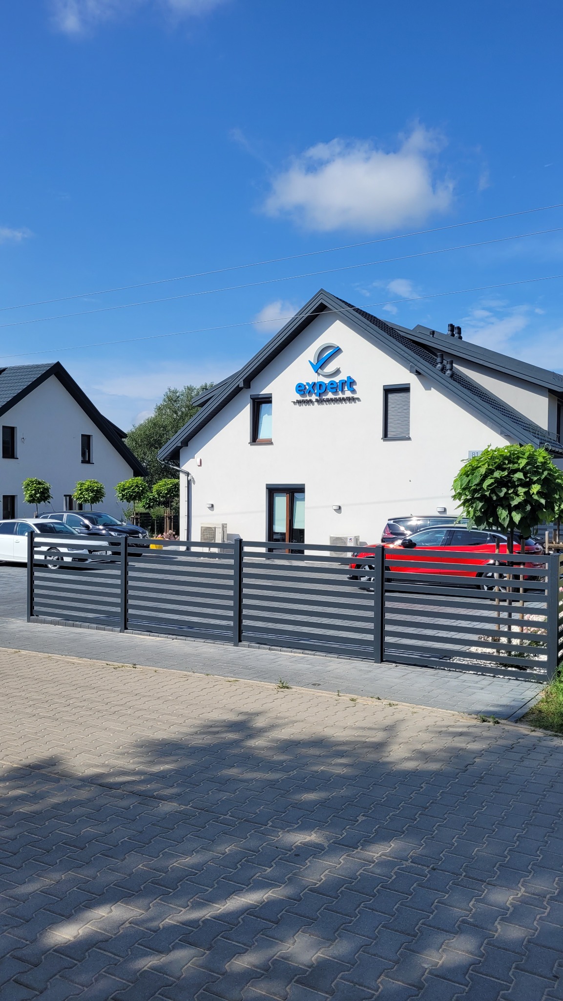 Dwukondygnacyjny budynek biurowy z szarą dachówką i białymi ścianami, z niebieskim logo 'expert' nad wejściem, ogrodzony szarą, poziomą metalową barierką, z zaparkowanymi samochodami i drzewami w tle.