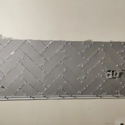 Ściana w trakcie układania szarych, podłużnych płytek w jodełkę, z widocznymi krzyżykami dystansowymi i puszkami elektrycznymi.