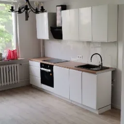 Nowoczesna kuchnia z białymi szafkami, drewnianym blatem, czarną armaturą i marmurową ścianą nad blatem, widziana z perspektywy podłogi z paneli.