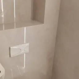 Biała toaleta z deską wolnoopadającą i przyciskiem spłukującym na ścianie wyłożonej beżowymi płytkami imitującymi marmur, oświetlenie LED w fugach, widoczny fragment szczotki do WC