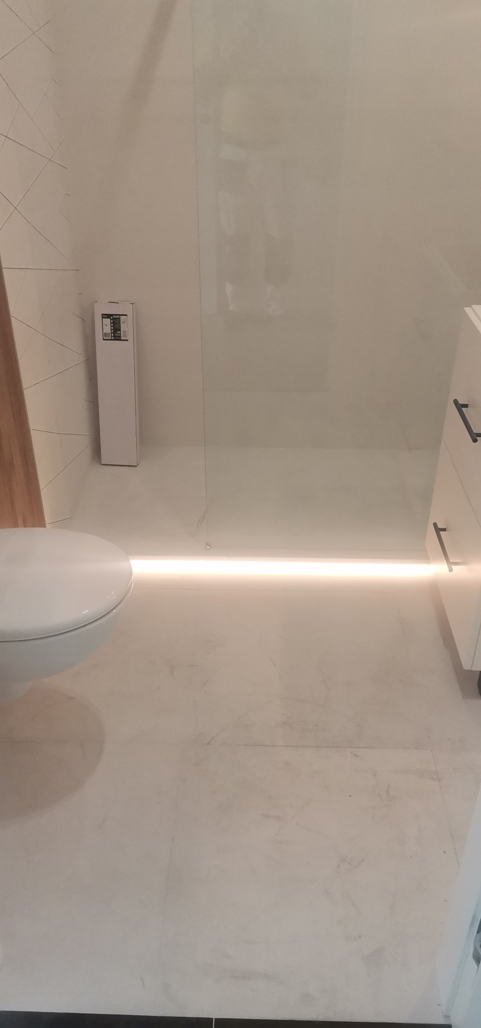 Nowoczesna łazienka z podświetlaną podłogą i minimalistyczną toaletą oraz kabiną prysznicową ze szkła, widoczna paczka produktu przy ścianie