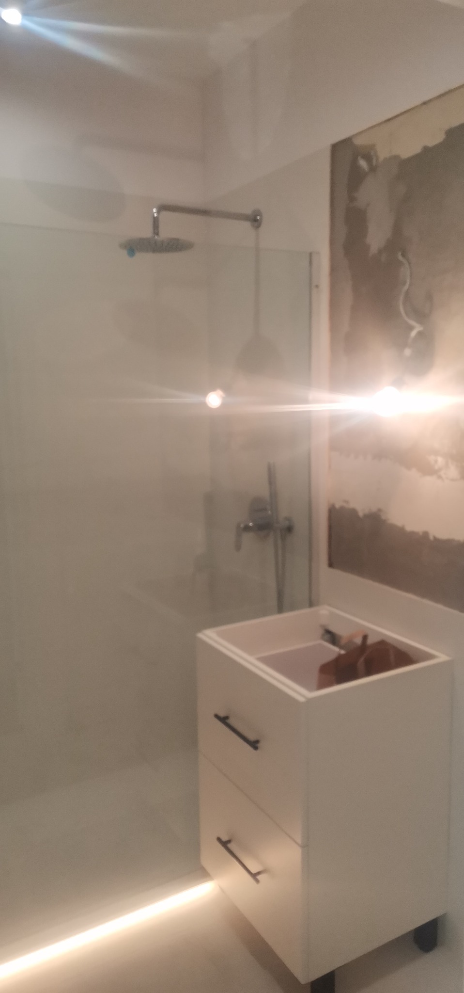 Nowa kabina prysznicowa z deszczownicą i białą szafką z dwoma szufladami, widoczne ślady po remoncie na ścianie.