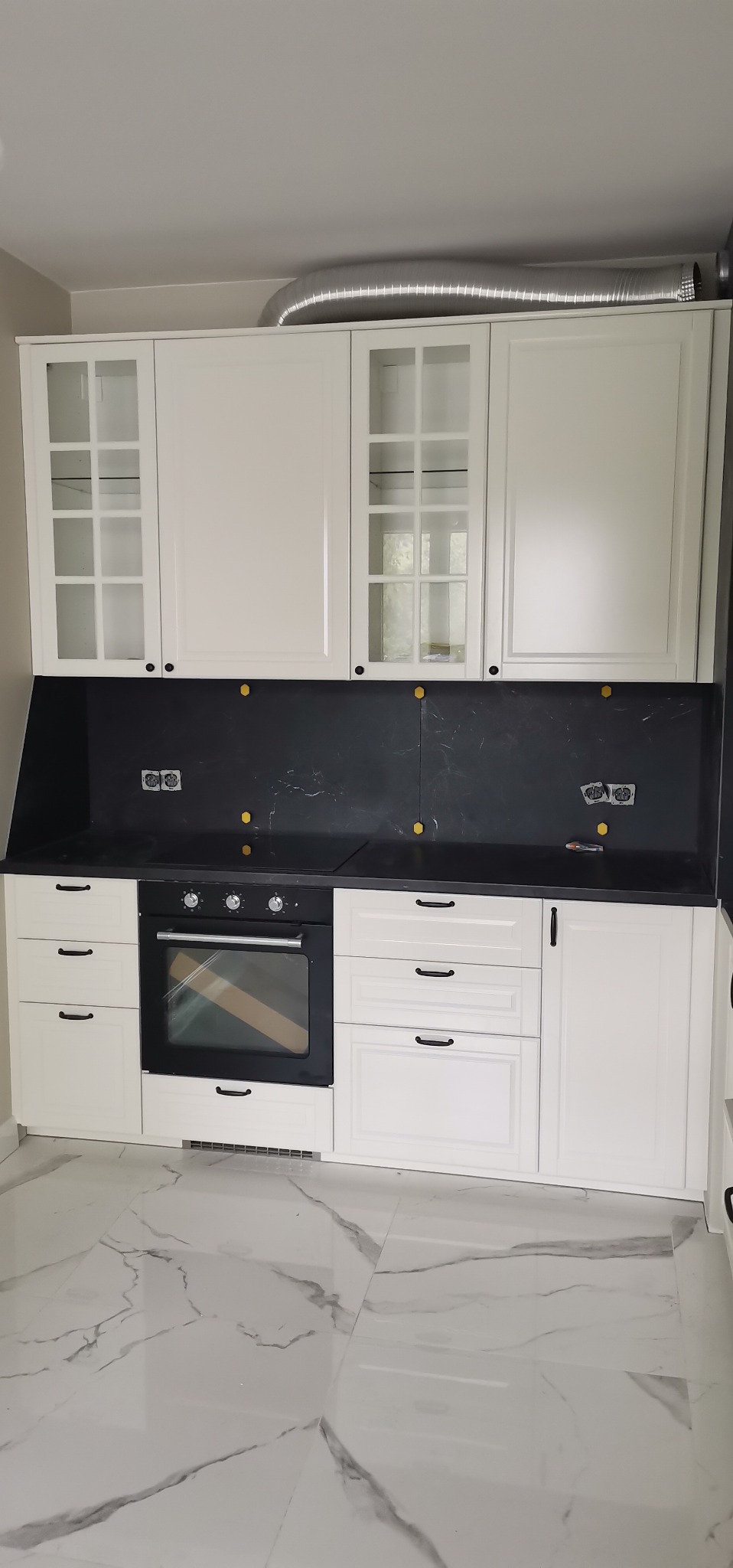 Biała zabudowa kuchenna z czarnym panelem nad blatem, widoczne gniazdka elektryczne i fragment wentylacji.
