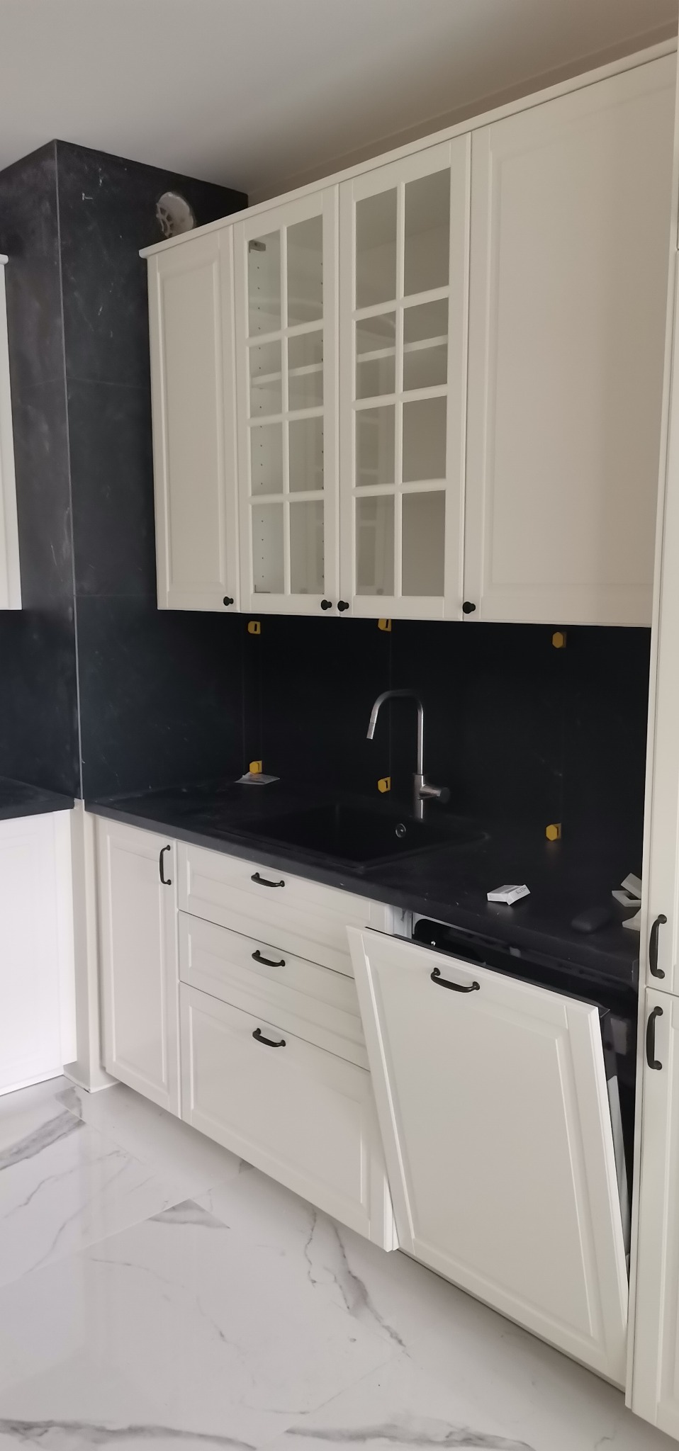 Nowoczesna kuchnia z białymi szafkami, czarnym blatem i marmurową podłogą w trakcie montażu, widoczne żółte podkładki poziomujące na blacie.