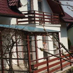 Dwupiętrowy dom z białymi ścianami, brązowym dachem z dachówki i balkonem z drewnianą balustradą, widoczna antena satelitarna i elementy ogrodu z drewnianym obramowaniem rabatki.