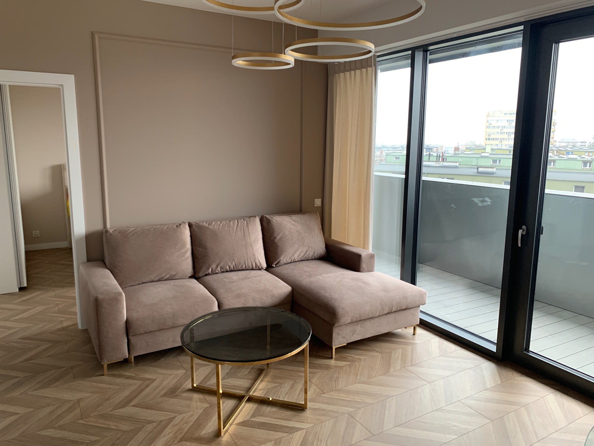 Jasne wnętrze salonu z beżową sofą, okrągłym stolikiem kawowym ze złotymi nogami, parkietem w jodełkę i dużym oknem balkonowym z widokiem na miasto.