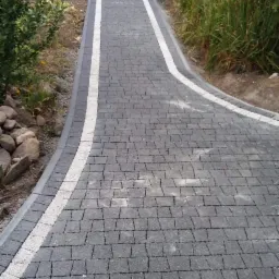 Ułożona ścieżka z ciemnoszarej kostki brukowej z białymi pasami po bokach, prowadząca przez bramę z cegły, z łopatą opartą o jeden ze słupków.