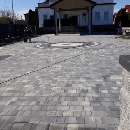 Błażej Świerczek - Szara kostka brukowa ułożona na posesji przed domem, pracownik czyści powierzchnię myjką ciśnieniową, widoczne elementy dekoracyjne z kostki w formie okręgów, fragment murku z kostki po prawej...