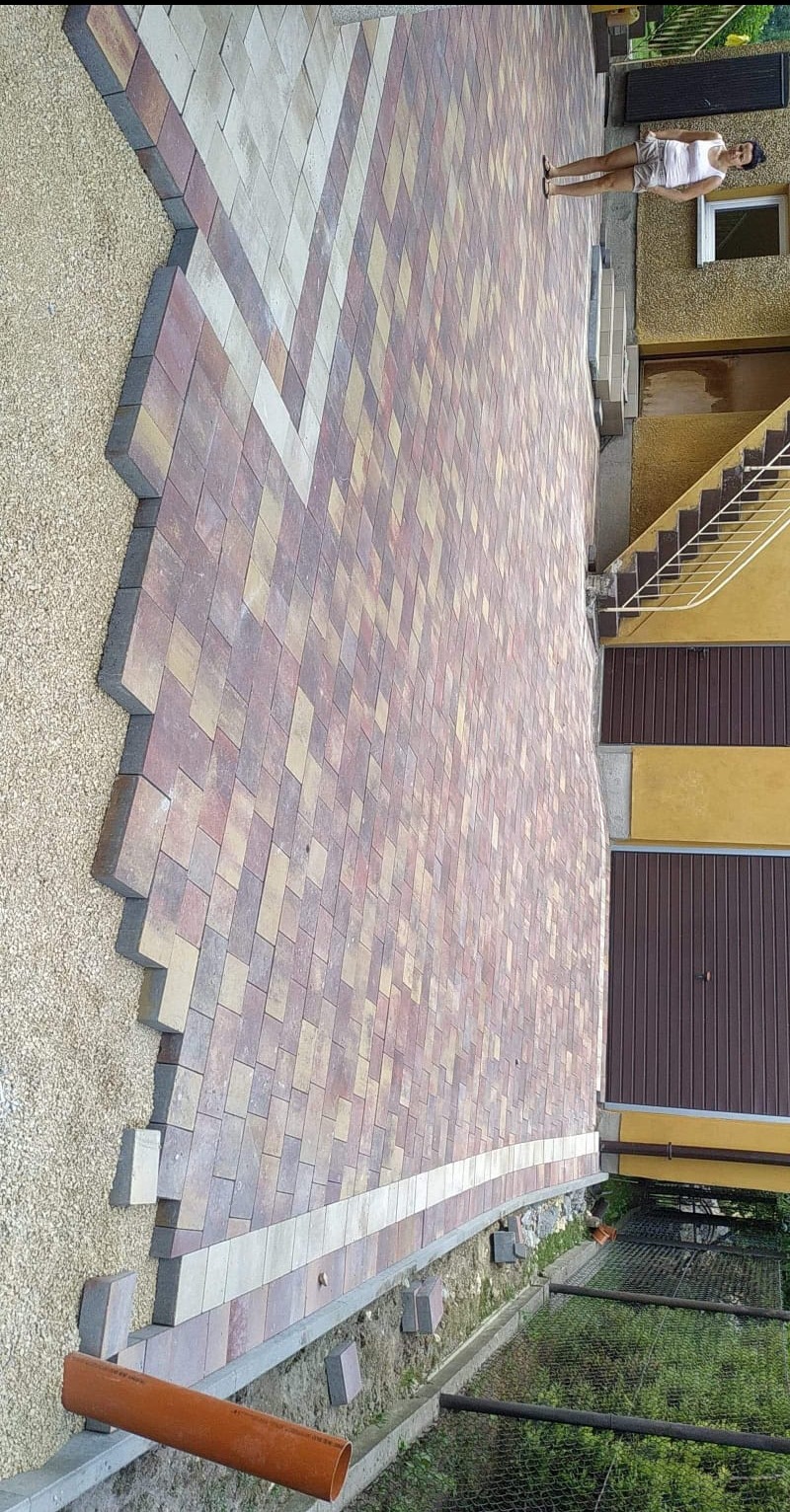 Fragment nowo ułożonej, kolorowej kostki brukowej w geometryczny wzór, przylegającej do budynku z brązową bramą garażową; widoczny pomarańczowy rurociąg i obszar przygotowany pod dalsze prace.